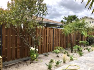 Backyard with a tall wooden fence, a flowering bush, and tropical plants under a cloudy sky; a house with a tiled roof is visible in the background.